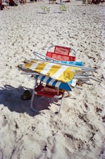 A red beach chair is placed on sandy terrain, stacked with multiple folded beach umbrellas featuring striped patterns in blue, yellow, and white. In the background, other beachgoers are visible along with a few green chairs scattered across the sand.