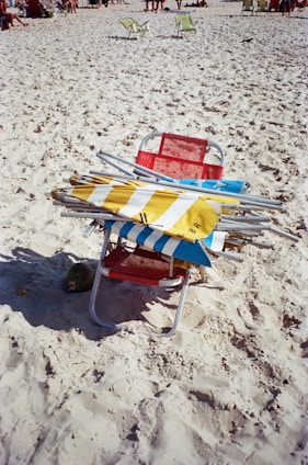 A red beach chair is placed on sandy terrain, stacked with multiple folded beach umbrellas featuring striped patterns in blue, yellow, and white. In the background, other beachgoers are visible along with a few green chairs scattered across the sand.
