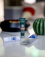 A collection of skincare products are arranged on a glossy surface. The central focus is a green tube from L'Occitane with intricate labeling. Surrounding it are a white cylindrical container and a gray tube with blue accents. In the background, a decorative green and white ceramic object and a blurred, abstract pattern add to the aesthetic.