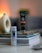 A tube of L'Occitane hair product labeled 'Intensive Repair' stands upright on a stack of magazines and books. The background is softly blurred, featuring a cream-colored decorative object with a round opening. The setting appears indoors with warm ambient lighting.