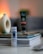A tube of L'Occitane hair product labeled 'Intensive Repair' stands upright on a stack of magazines and books. The background is softly blurred, featuring a cream-colored decorative object with a round opening. The setting appears indoors with warm ambient lighting.
