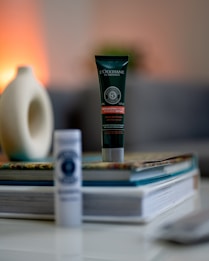 A tube of L'Occitane hair product labeled 'Intensive Repair' stands upright on a stack of magazines and books. The background is softly blurred, featuring a cream-colored decorative object with a round opening. The setting appears indoors with warm ambient lighting.