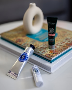 Three cosmetic products are arranged on a table. A silver tube of hand cream lies horizontally in the foreground next to a small white bottle. Behind them, a dark green tube stands upright on a colorful book. In the background, a white abstract ceramic vase sits atop the book.