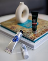 Three cosmetic products are arranged on a table. A silver tube of hand cream lies horizontally in the foreground next to a small white bottle. Behind them, a dark green tube stands upright on a colorful book. In the background, a white abstract ceramic vase sits atop the book.