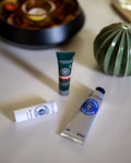 A neatly arranged collection of cosmetic products from L'Occitane includes a tube of hand cream, a lip balm stick, and a small facial product, all placed on a smooth, light-colored surface. A decorative green vase is visible in the background alongside a shallow bowl with decorative items.