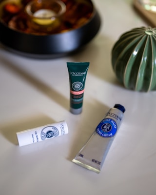 A neatly arranged collection of cosmetic products from L'Occitane includes a tube of hand cream, a lip balm stick, and a small facial product, all placed on a smooth, light-colored surface. A decorative green vase is visible in the background alongside a shallow bowl with decorative items.