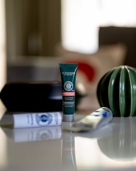A tube of L'Occitane product is centered on a sleek reflective surface. Surrounding it are other skincare tubes and a decorative vase with green and white stripes. The background is softly blurred, creating a serene and elegant atmosphere.