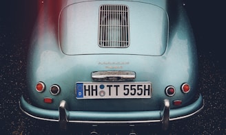 A classic car with a sleek, teal body displaying raindrops. The rear view features distinctive round tail lights and a chrome bumper. The license plate reads 'HH TT 555H,' and the make of the car is prominently displayed above the license plate.