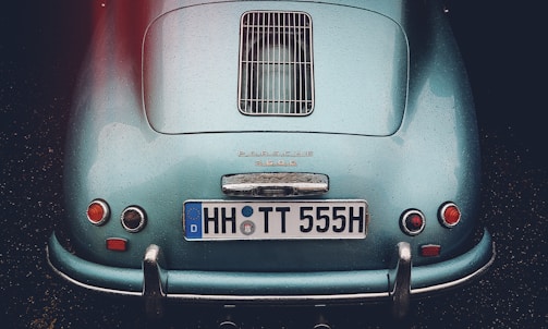 A classic car with a sleek, teal body displaying raindrops. The rear view features distinctive round tail lights and a chrome bumper. The license plate reads 'HH TT 555H,' and the make of the car is prominently displayed above the license plate.
