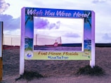 A decorative photo frame is placed on a sandy beach, with a sign reading 'Wish You Were Here!' and 'Fort Pierce, Florida #LoveTheFort'. The frame is bordered by illustrations of palm leaves and ocean scenes, set against a backdrop featuring a cloudy sky and a red netting fence.