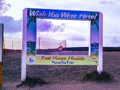 A decorative photo frame is placed on a sandy beach, with a sign reading 'Wish You Were Here!' and 'Fort Pierce, Florida #LoveTheFort'. The frame is bordered by illustrations of palm leaves and ocean scenes, set against a backdrop featuring a cloudy sky and a red netting fence.