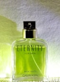 A clear glass bottle of Calvin Klein's 'Eternity for Men' Eau de Toilette is centered against a white and yellow gradient background. The bottle contains a green liquid and has a silver cap with a black spray nozzle. The text 'ETERNITY FOR MEN' is prominently displayed on the front of the bottle along with 'EAU DE TOILETTE' and 'CALVIN KLEIN' at the bottom.