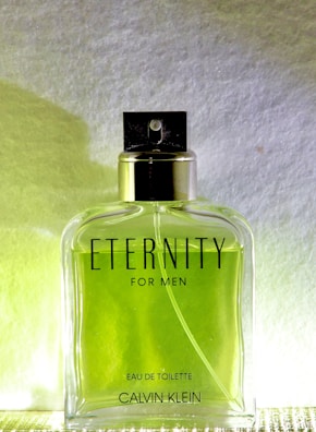 A clear glass bottle of Calvin Klein's 'Eternity for Men' Eau de Toilette is centered against a white and yellow gradient background. The bottle contains a green liquid and has a silver cap with a black spray nozzle. The text 'ETERNITY FOR MEN' is prominently displayed on the front of the bottle along with 'EAU DE TOILETTE' and 'CALVIN KLEIN' at the bottom.