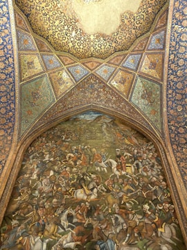 An intricately detailed mural depicts a historical battle scene with numerous figures on horseback, engaged in combat, featuring traditional attire and weaponry. Above the scene, an ornately decorated arch with geometric patterns and floral motifs showcases vibrant colors and elaborate designs that suggest a rich cultural heritage.