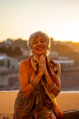 An elderly woman with short hair is smiling warmly while clasping her hands together. She is adorned in colorful clothing and bracelets, sitting on a rooftop with a blurred cityscape and glowing sunset in the background.