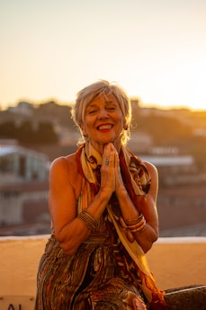 An elderly woman with short hair is smiling warmly while clasping her hands together. She is adorned in colorful clothing and bracelets, sitting on a rooftop with a blurred cityscape and glowing sunset in the background.