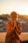 An elderly woman with short hair is smiling warmly while clasping her hands together. She is adorned in colorful clothing and bracelets, sitting on a rooftop with a blurred cityscape and glowing sunset in the background.