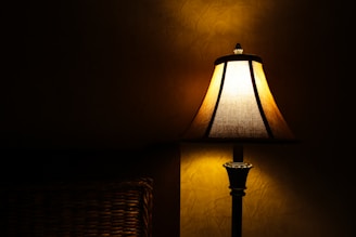A warmly lit table lamp with a fabric shade casts a soft glow against a textured wall. The lamp's base is dark-colored, adding contrast to its soft light.