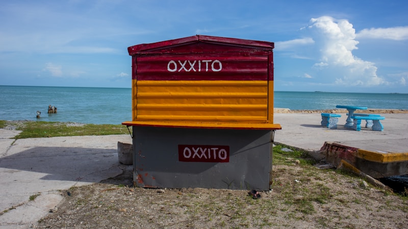A colorful kiosk with the word 'OXXITO' is situated on a concrete platform near a beach. The kiosk has a red and yellow striped top and a gray base. In the background, there's a calm ocean under a partly cloudy sky. To the right, there are turquoise-painted benches and tables.