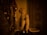 A detailed sculpture of a monkey sits on a shelf, lit by warm lighting that casts dramatic shadows. Nearby are ornate candlesticks and a reflective surface that adds to the ambiance. The setting has an old-world, vintage feel, with intricate detailing on the door and surrounding decor.