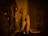 A detailed sculpture of a monkey sits on a shelf, lit by warm lighting that casts dramatic shadows. Nearby are ornate candlesticks and a reflective surface that adds to the ambiance. The setting has an old-world, vintage feel, with intricate detailing on the door and surrounding decor.