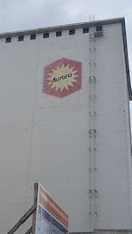 A large industrial building facade with a hexagonal red and yellow logo displaying the word 'Aurora'. The building has a tall vertical ladder on its side, reaching up to the roof. Below, there is a construction sign with text in German against a cloudy sky backdrop.