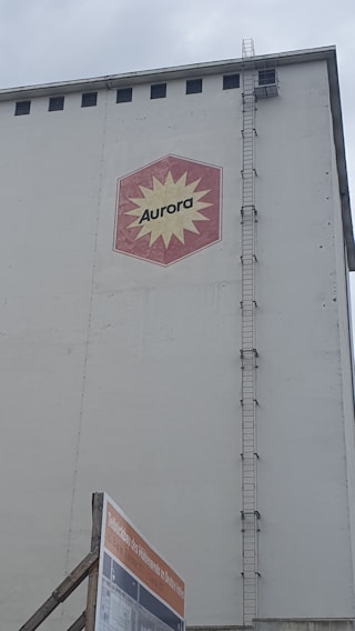 A large industrial building facade with a hexagonal red and yellow logo displaying the word 'Aurora'. The building has a tall vertical ladder on its side, reaching up to the roof. Below, there is a construction sign with text in German against a cloudy sky backdrop.