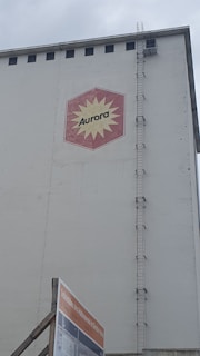 A large industrial building facade with a hexagonal red and yellow logo displaying the word 'Aurora'. The building has a tall vertical ladder on its side, reaching up to the roof. Below, there is a construction sign with text in German against a cloudy sky backdrop.