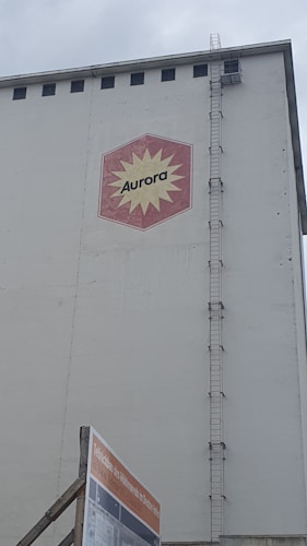 A large industrial building facade with a hexagonal red and yellow logo displaying the word 'Aurora'. The building has a tall vertical ladder on its side, reaching up to the roof. Below, there is a construction sign with text in German against a cloudy sky backdrop.