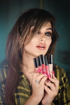 A woman with styled, medium-length hair holds several liquid lipsticks in front of her face. She has dramatic eye makeup and glossy lips. She is wearing a patterned shirt and a large ring on her finger.