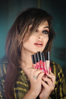 A woman with styled, medium-length hair holds several liquid lipsticks in front of her face. She has dramatic eye makeup and glossy lips. She is wearing a patterned shirt and a large ring on her finger.