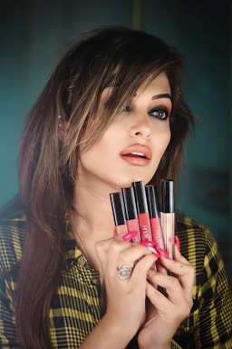 A woman with styled, medium-length hair holds several liquid lipsticks in front of her face. She has dramatic eye makeup and glossy lips. She is wearing a patterned shirt and a large ring on her finger.