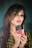 A woman with styled, medium-length hair holds several liquid lipsticks in front of her face. She has dramatic eye makeup and glossy lips. She is wearing a patterned shirt and a large ring on her finger.