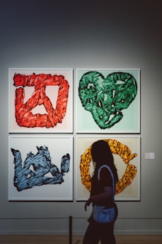 Four framed abstract artworks are displayed on a gallery wall, each with a distinct color theme: red, green, blue, and yellow. The art features intricate, textured patterns and shapes resembling letters or symbols. A person walks in front of the artworks, silhouetted against the light gray wall.