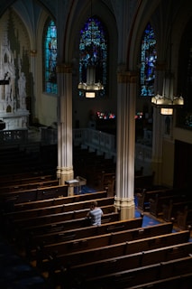 An interior of a church featuring wooden pews, ornate columns, and stained glass windows casting colorful light. A solitary person sits in contemplation, wearing a light-colored top. Soft light illuminates the area from overhead fixtures.