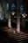 An interior of a church featuring wooden pews, ornate columns, and stained glass windows casting colorful light. A solitary person sits in contemplation, wearing a light-colored top. Soft light illuminates the area from overhead fixtures.