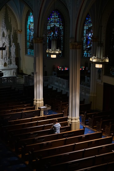 An interior of a church featuring wooden pews, ornate columns, and stained glass windows casting colorful light. A solitary person sits in contemplation, wearing a light-colored top. Soft light illuminates the area from overhead fixtures.