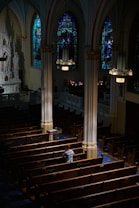 An interior of a church featuring wooden pews, ornate columns, and stained glass windows casting colorful light. A solitary person sits in contemplation, wearing a light-colored top. Soft light illuminates the area from overhead fixtures.