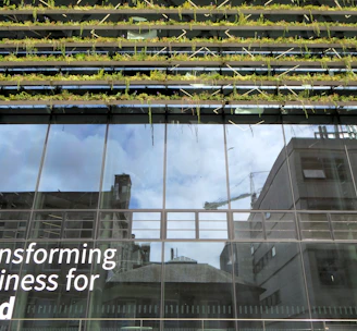 Glass-fronted building with reflections of the sky and nearby structures. Rows of greenery are integrated into the design above the windows. Large text on the building's facade reads 'Transforming Business for Good.'