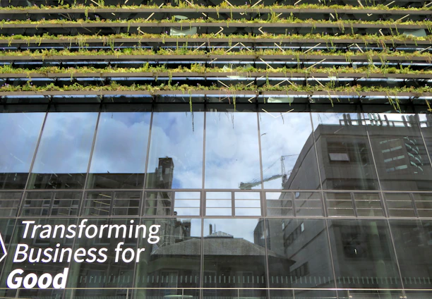 Glass-fronted building with reflections of the sky and nearby structures. Rows of greenery are integrated into the design above the windows. Large text on the building's facade reads 'Transforming Business for Good.'