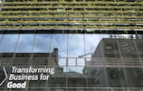 Glass-fronted building with reflections of the sky and nearby structures. Rows of greenery are integrated into the design above the windows. Large text on the building's facade reads 'Transforming Business for Good.'