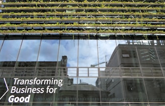 Glass-fronted building with reflections of the sky and nearby structures. Rows of greenery are integrated into the design above the windows. Large text on the building's facade reads 'Transforming Business for Good.'