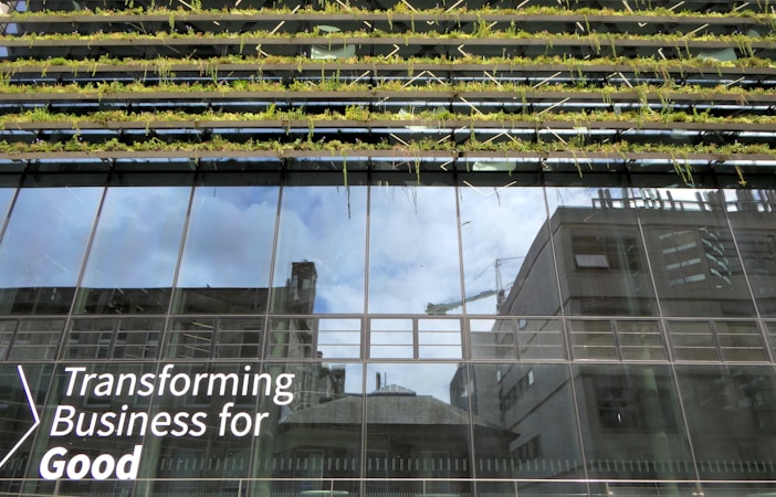 Glass-fronted building with reflections of the sky and nearby structures. Rows of greenery are integrated into the design above the windows. Large text on the building's facade reads 'Transforming Business for Good.'