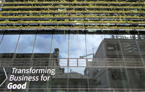 Glass-fronted building with reflections of the sky and nearby structures. Rows of greenery are integrated into the design above the windows. Large text on the building's facade reads 'Transforming Business for Good.'