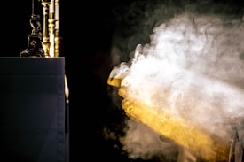 A waft of smoke or mist, illuminated by a gentle light, flows across a dark background. To the left, an altar or table is partially visible, draped in a white cloth. Several ornate golden candlesticks are positioned prominently, adding a sense of opulence.
