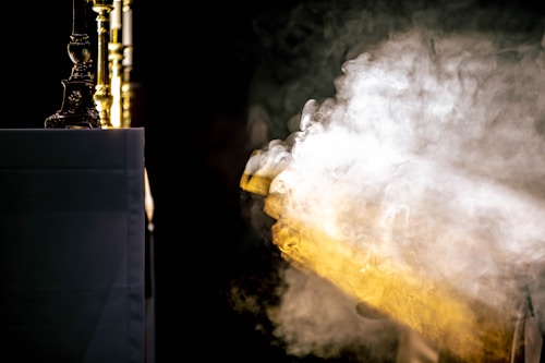 A waft of smoke or mist, illuminated by a gentle light, flows across a dark background. To the left, an altar or table is partially visible, draped in a white cloth. Several ornate golden candlesticks are positioned prominently, adding a sense of opulence.