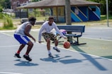 Two people are playing basketball on an outdoor court. One player is dribbling the ball while the other plays defense. The court is surrounded by grass and there is a bench with clothes and a bottle next to it. A building is visible in the background.