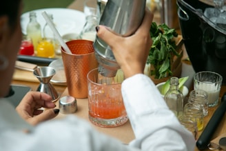 A person is in the process of mixing a cocktail, using a cocktail shaker and pouring the mixture into a clear glass. Various cocktail-making items are spread on the table, including a jigger, strainer, and multiple small bottles with colorful liquids. Fresh mint leaves are also visible, along with an ice bucket and a copper mug.