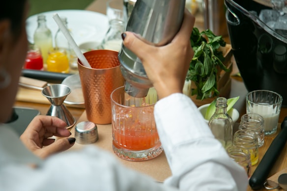 A person is in the process of mixing a cocktail, using a cocktail shaker and pouring the mixture into a clear glass. Various cocktail-making items are spread on the table, including a jigger, strainer, and multiple small bottles with colorful liquids. Fresh mint leaves are also visible, along with an ice bucket and a copper mug.