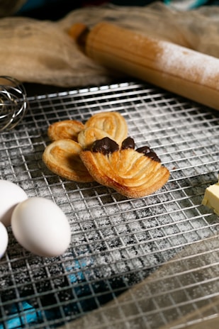 A baking scene features flaky pastries with chocolate, resting on a cooling rack dusted with flour. Nearby, fresh eggs, a block of butter, a whisk, and a wooden rolling pin are visible, suggesting a preparation setting. The overall atmosphere conveys a sense of home baking, warmth, and deliciousness.